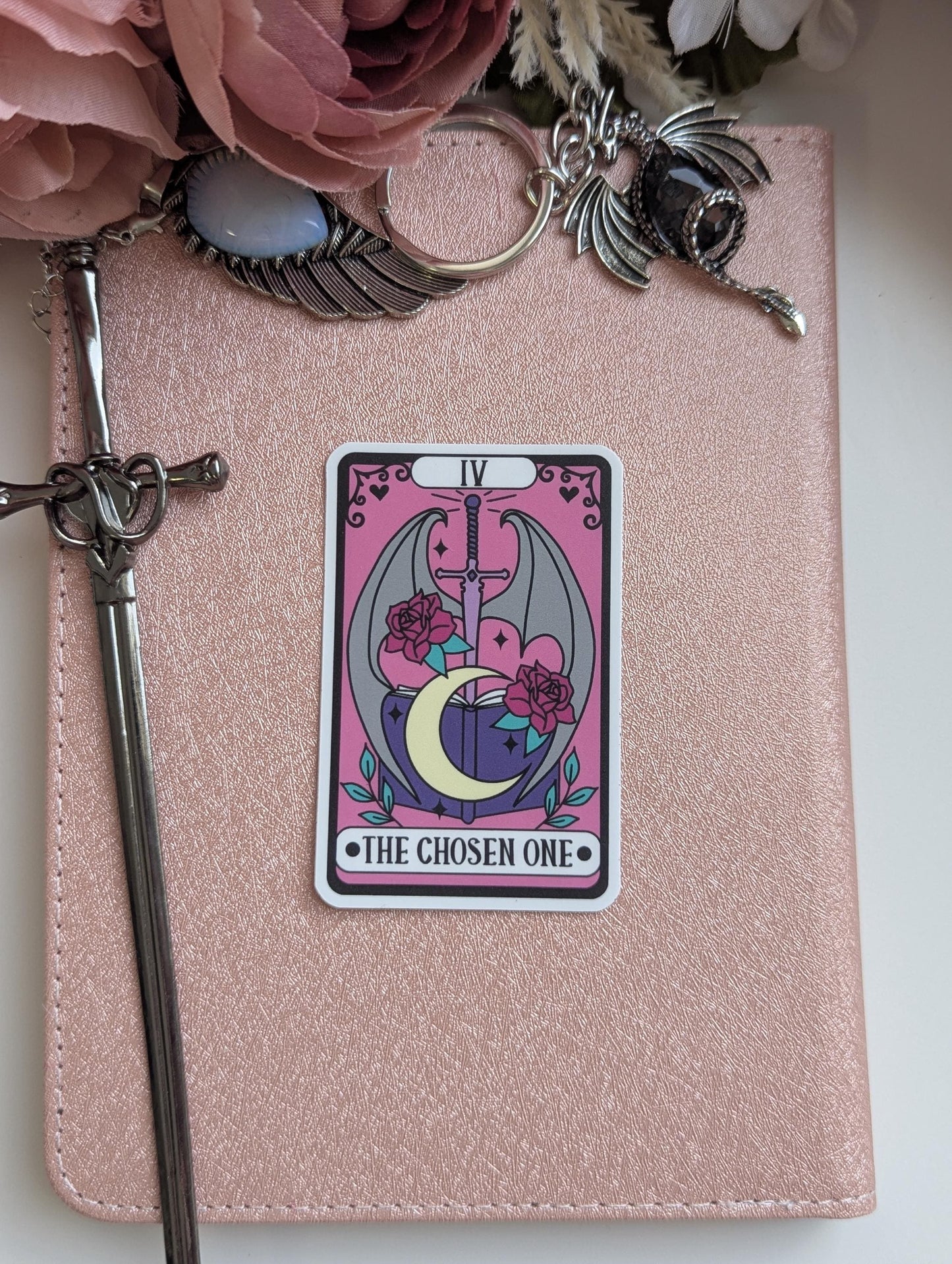 May include: A rectangular sticker featuring a tarot card design with a pink background, a central sword, a crescent moon, an open book, and two red roses. The text IV and THE CHOSEN ONE are printed on the sticker, which is placed on a rose gold notebook.