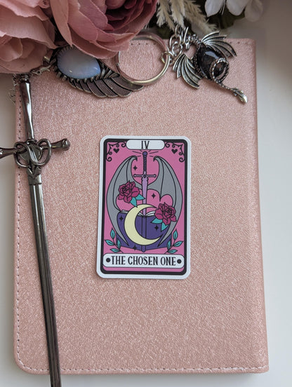 May include: A rectangular sticker featuring a tarot card design with a pink background, a central sword, a crescent moon, an open book, and two red roses. The text IV and THE CHOSEN ONE are printed on the sticker, which is placed on a rose gold notebook.