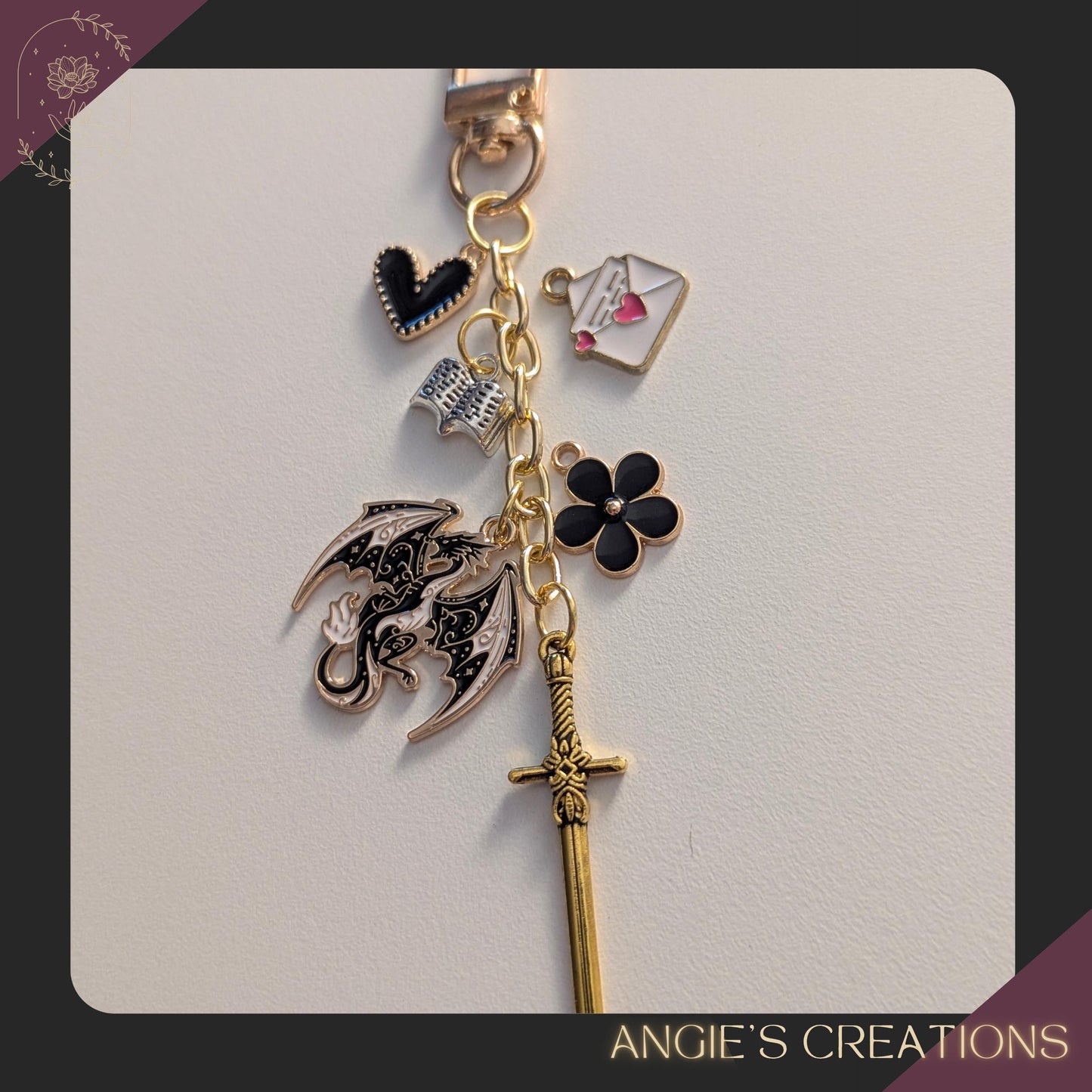 May include: A gold-coloured keychain with various charms. Charms include a black heart, an open book, an envelope with a heart, a black flower, a dragon, and a sword. The text "ANGIE'S CREATIONS" is at the bottom.