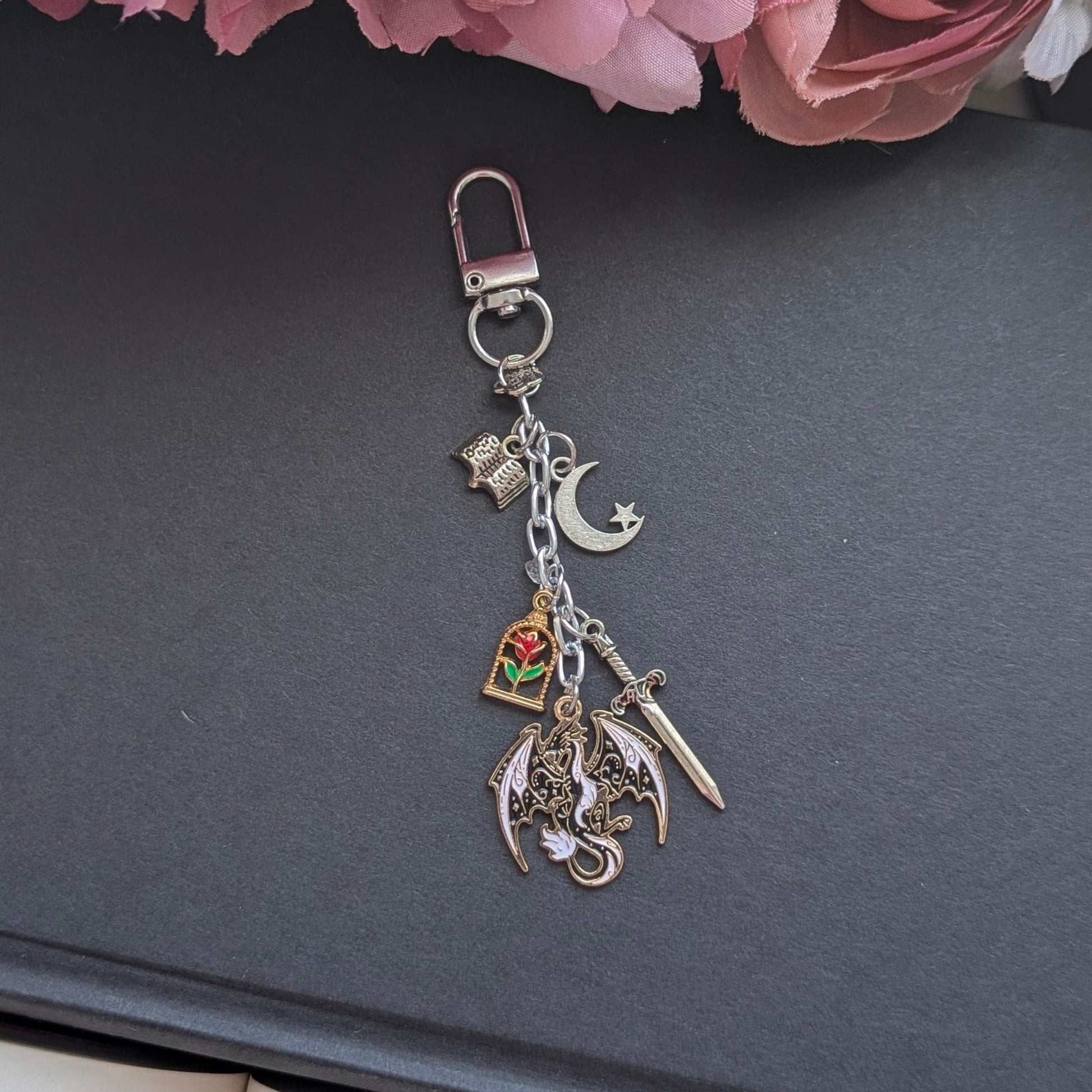 May include: Silver keychain featuring various charms: a dragon, a rose in a glass case, a crescent moon, a sword, and a book. The charms are suspended from a silver chain and clasp. Set against a dark grey backdrop.