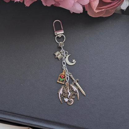 May include: Silver keychain featuring various charms: a dragon, a rose in a glass case, a crescent moon, a sword, and a book. The charms are suspended from a silver chain and clasp. Set against a dark grey backdrop.