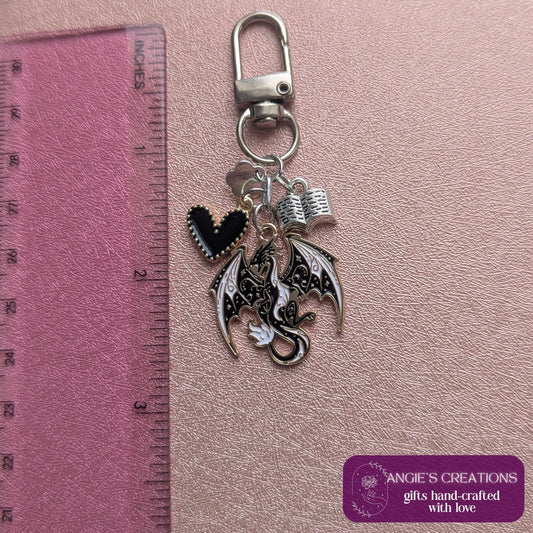 May include: A silver keychain with a lobster clasp, featuring charms of a black heart, a dragon with white and black wings, and an open book. The dragon charm is approximately 3.8 cm in length. The background is a pink surface.
