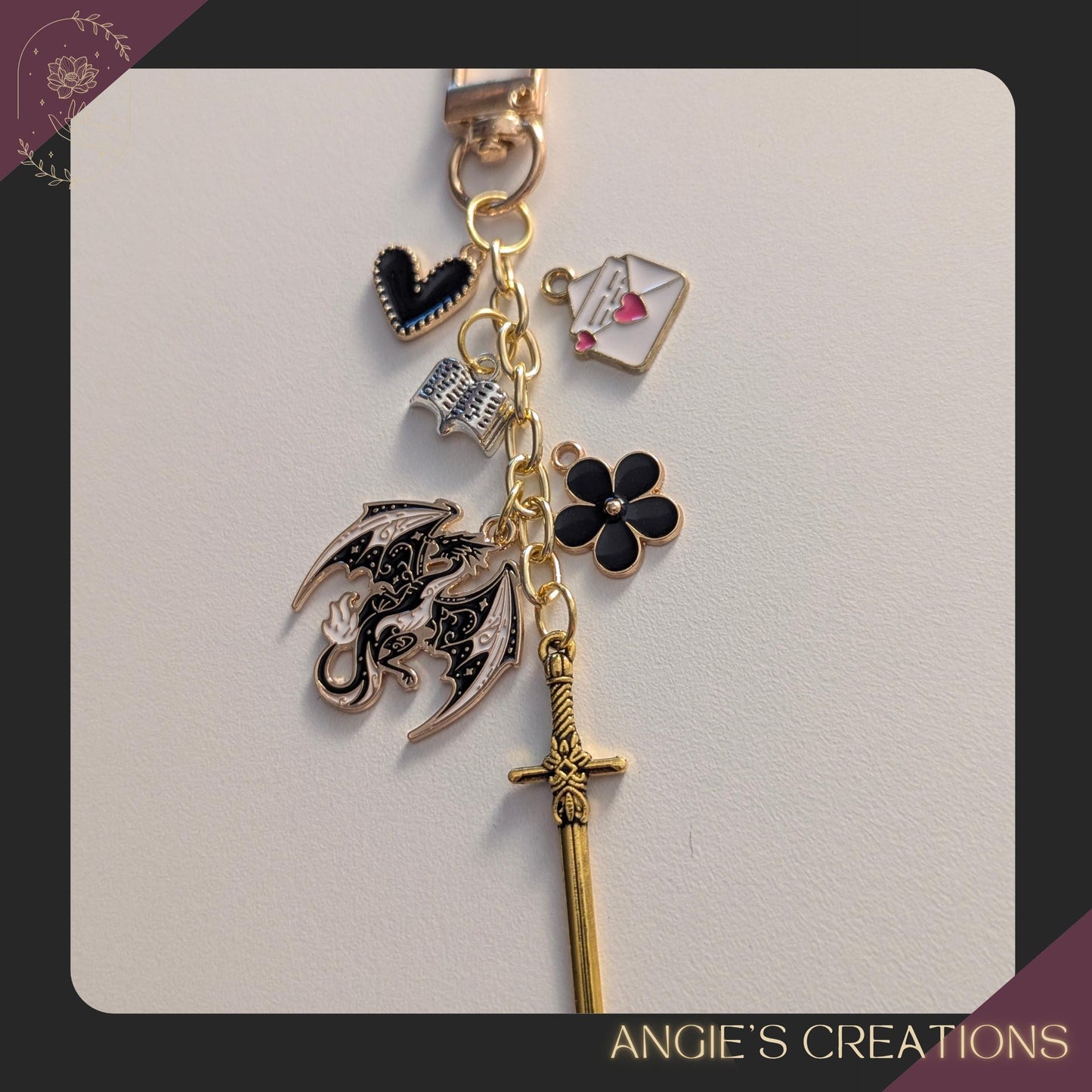 May include: A gold-coloured keychain with various charms. Charms include a black heart, an open book, an envelope with a heart, a black flower, a dragon, and a sword. The text "ANGIE'S CREATIONS" is at the bottom.