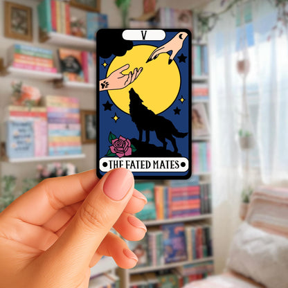 Angie's Creations handmade die-cut sticker featuring a dark tarot card style design labeled The Fated Mates card V, with two hands reaching toward each other over a full yellow moon with a black wolf silhouette howling below and a pink rose in the corner on a deep navy background, held against a blurred bookshelf background. Romantasy bookish tarot sticker designed in Ottawa Canada.