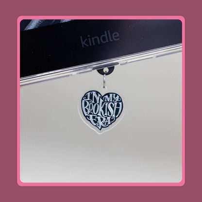 Black heart-shaped acrylic charm with white "In My Bookish Era" script lettering, sold as a matching Kindle grip and phone dust plug set.