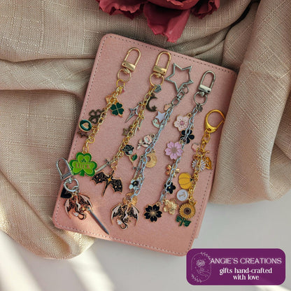 May include: Several handcrafted keychains with charms such as a four-leaf clover, a star, flowers, and a dragon. The keychains are displayed on a pink notebook. The image includes the text "ANGIE'S CREATIONS gifts hand-crafted with love."