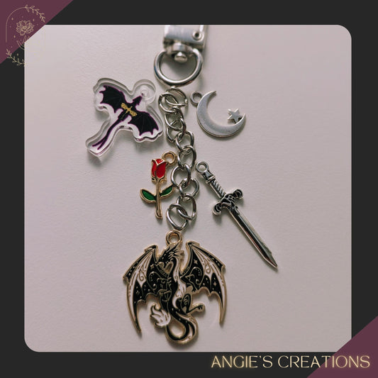 A handmade Kindle charm chain featuring a purple dragon acrylic charm, silver sword, crescent moon, and red rose charms on a silver chain. Fantasy romantasy accessory for readers and book lovers.