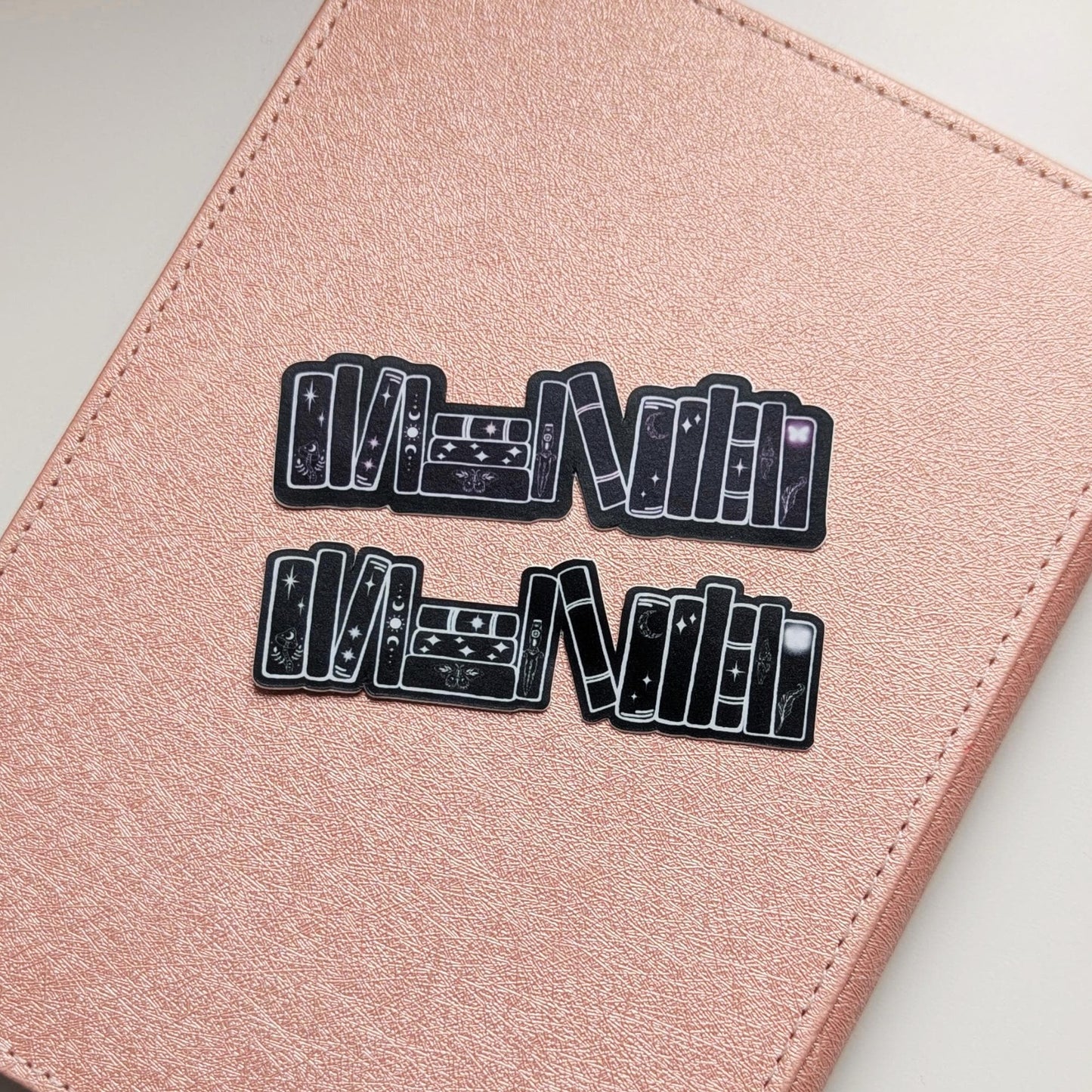 May include: Two black stickers with a stack of books design, featuring celestial elements like moons and stars. The stickers are placed on a pink leather-like surface, possibly a notebook or journal.