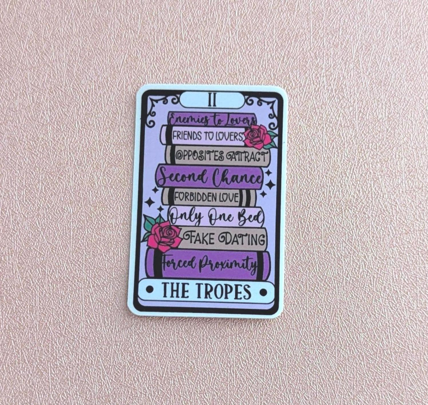 May include: A rectangular sticker designed like a tarot card featuring a stack of books in shades of purple and grey. The books are labelled with text including Enemies to Lovers, Friends to Lovers, Opposites Attract, Second Chance, Forbidden Love, Only One Bed, Fake Dating, and Forced Proximity, with pink roses and the text THE TROPES.