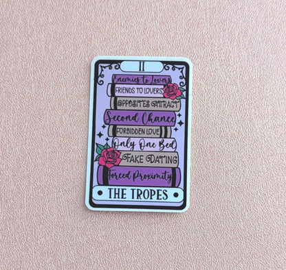 May include: A rectangular sticker designed like a tarot card featuring a stack of books in shades of purple and grey. The books are labelled with text including Enemies to Lovers, Friends to Lovers, Opposites Attract, Second Chance, Forbidden Love, Only One Bed, Fake Dating, and Forced Proximity, with pink roses and the text THE TROPES.