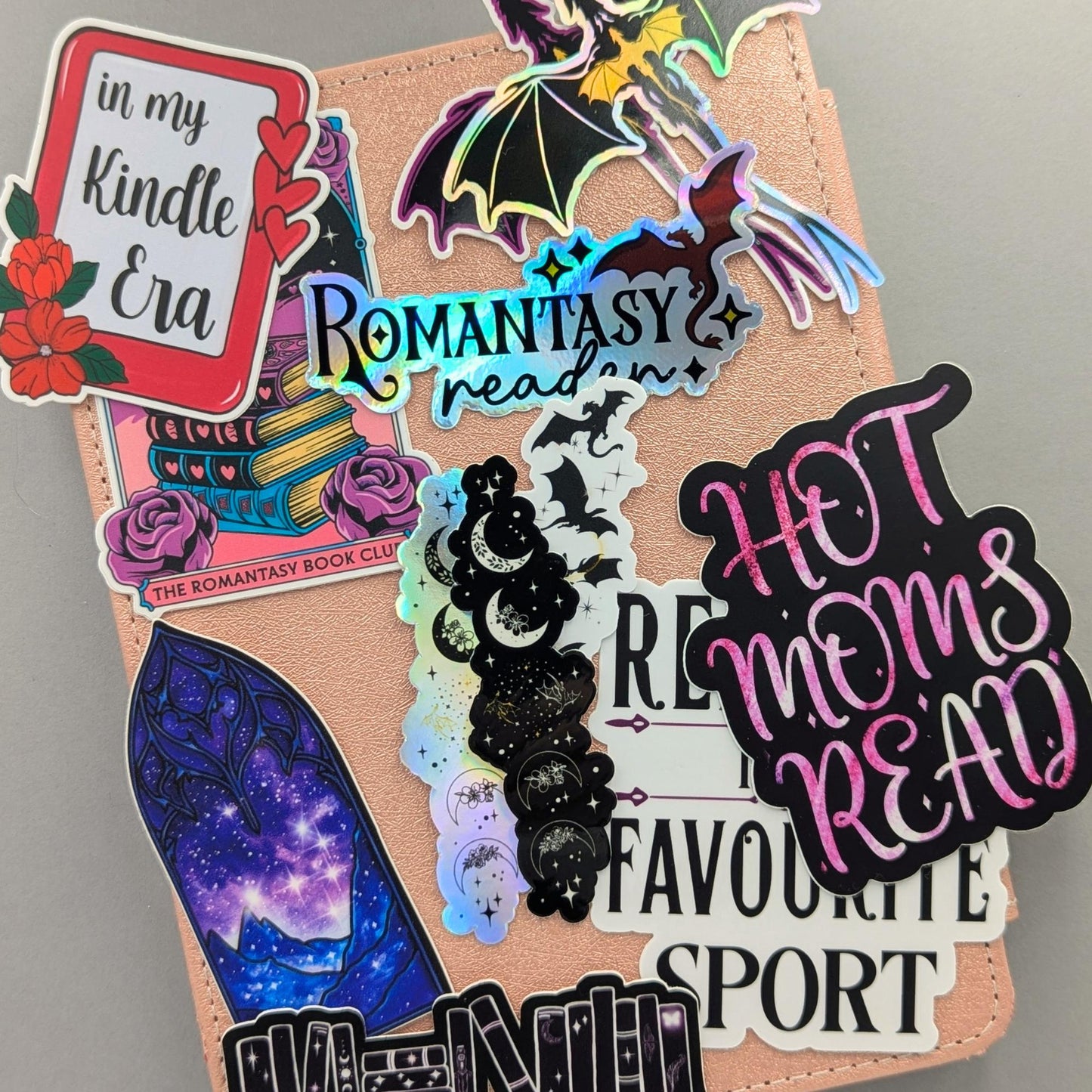 Collection of new romantasy and bookish waterproof dishwasher safe stickers displayed on rose gold Kindle eReader case, featuring holographic silver dragon sticker Romantasy Reader holographic text sticker Hot Moms Read foil effect sticker celestial moon phases sticker and fantasy inspired designs, ACOTAR Fourth Wing bookish gift ideas designed and manufactured in Canada by Angie's Creations