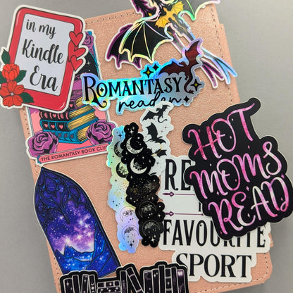 Collection of new romantasy and bookish waterproof dishwasher safe stickers displayed on rose gold Kindle eReader case, featuring holographic silver dragon sticker Romantasy Reader holographic text sticker Hot Moms Read foil effect sticker celestial moon phases sticker and fantasy inspired designs, ACOTAR Fourth Wing bookish gift ideas designed and manufactured in Canada by Angie's Creations