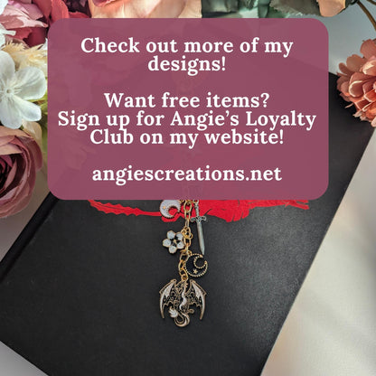 Hand assembled gold dragon charm keychain displayed on black hardcover romantasy book with pink and cream dried flowers, featuring celestial moon sword and floral charms, ACOTAR Fourth Wing inspired eReader Kindle accessory gift, promoting Angie's Loyalty Club at angiescreations.net designed in Ottawa Canada