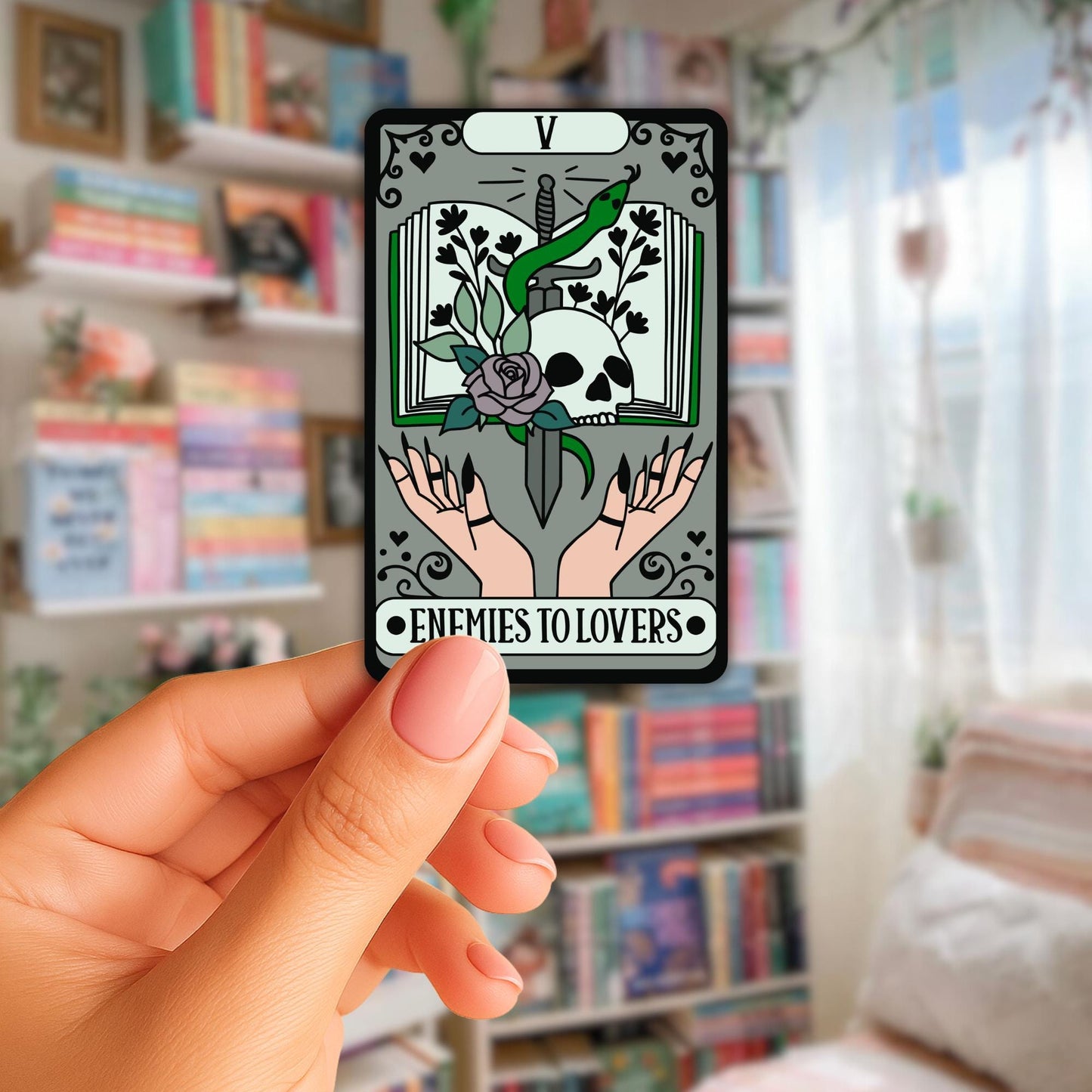 Angie's Creations handmade die-cut sticker featuring a dark tarot card style design labeled Enemies to Lovers card V, with two hands holding a sword wrapped in a green snake, a skull, dark rose, and botanical flowers on an open book with ornate grey card borders, held against a blurred bookshelf background. Bookish romantasy tarot sticker designed in Ottawa Canada.