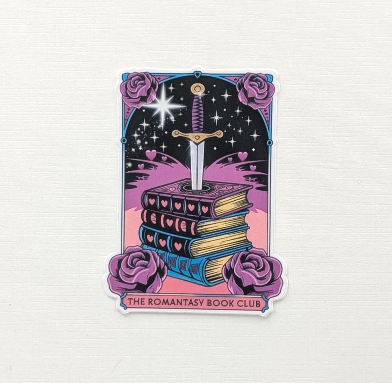 May include: A sticker depicting a stack of books pierced by a sword. The books are in pink, purple, and blue, with heart details. The background is black with stars and a starburst. The sticker has purple roses and the text "THE ROMANTASY BOOK CLUB".