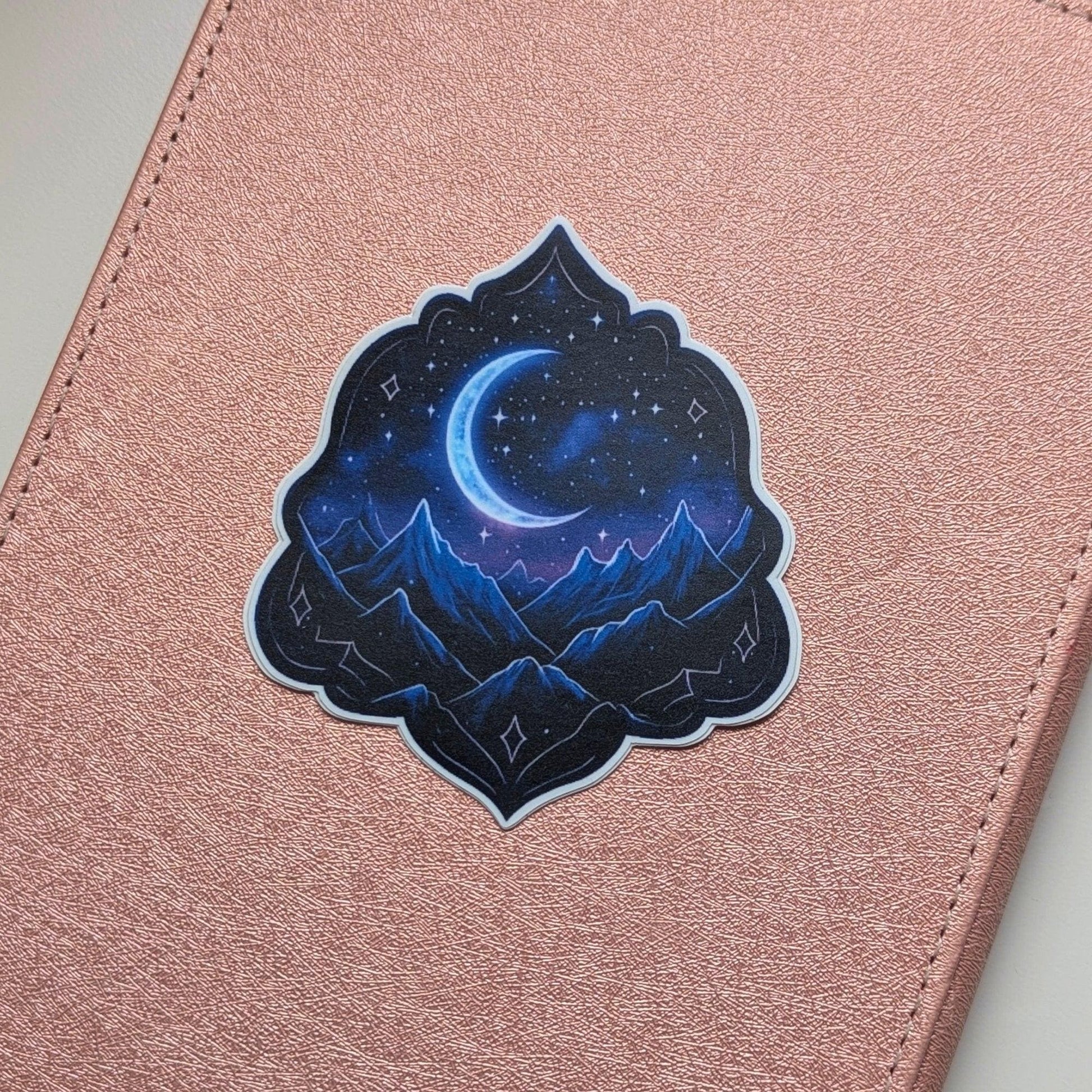 May include: A sticker depicting a crescent moon and stars above a mountain range. The sticker has a dark blue and purple colour scheme, with a decorative border. The sticker is placed on a pink, textured surface.