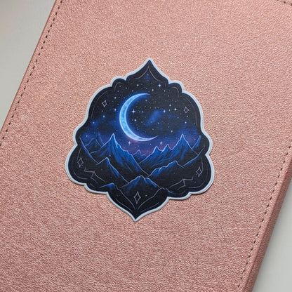 May include: A sticker depicting a crescent moon and stars above a mountain range. The sticker has a dark blue and purple colour scheme, with a decorative border. The sticker is placed on a pink, textured surface.