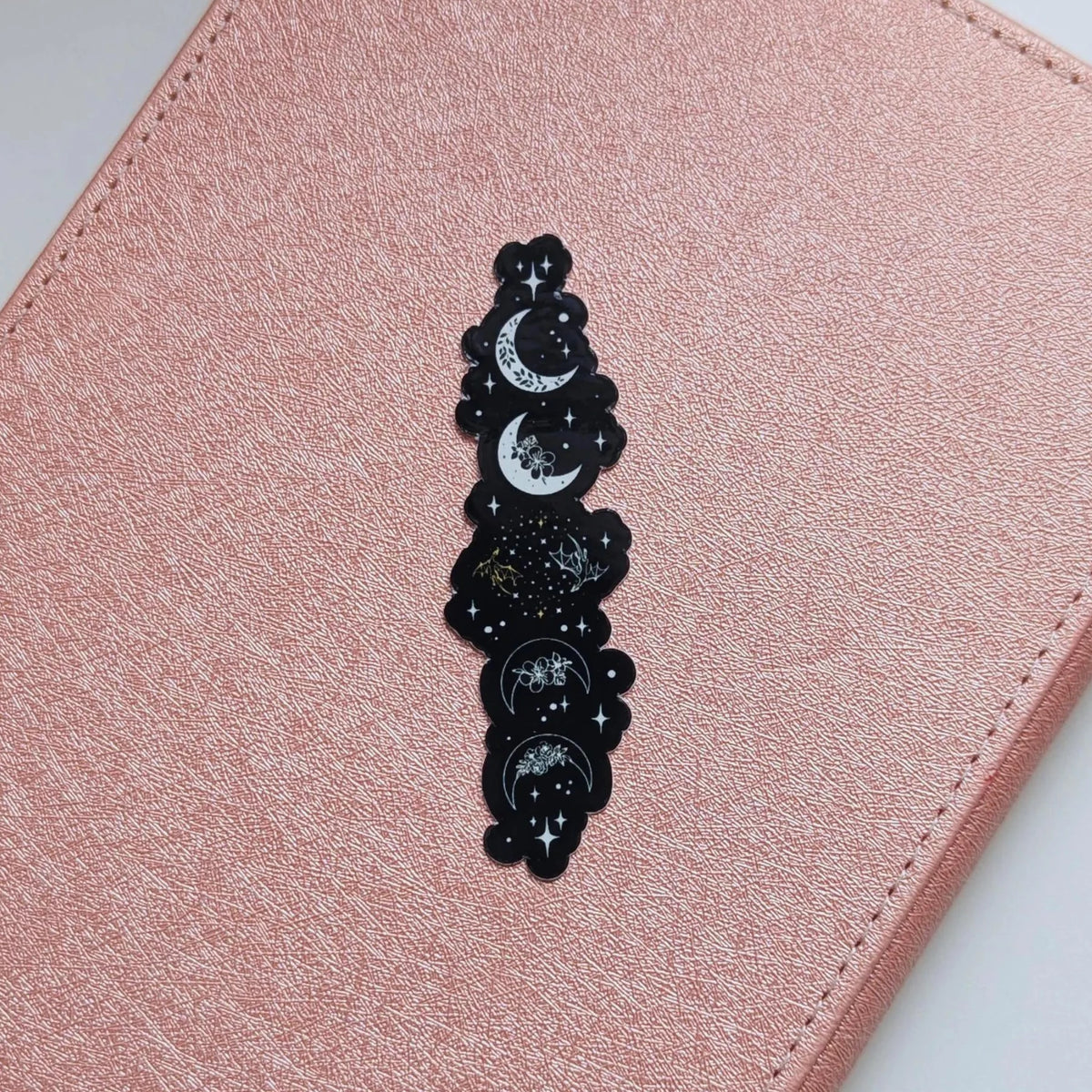 A close-up of a pink faux-leather surface with a vertical black enamel pin featuring moon and star motifs.
