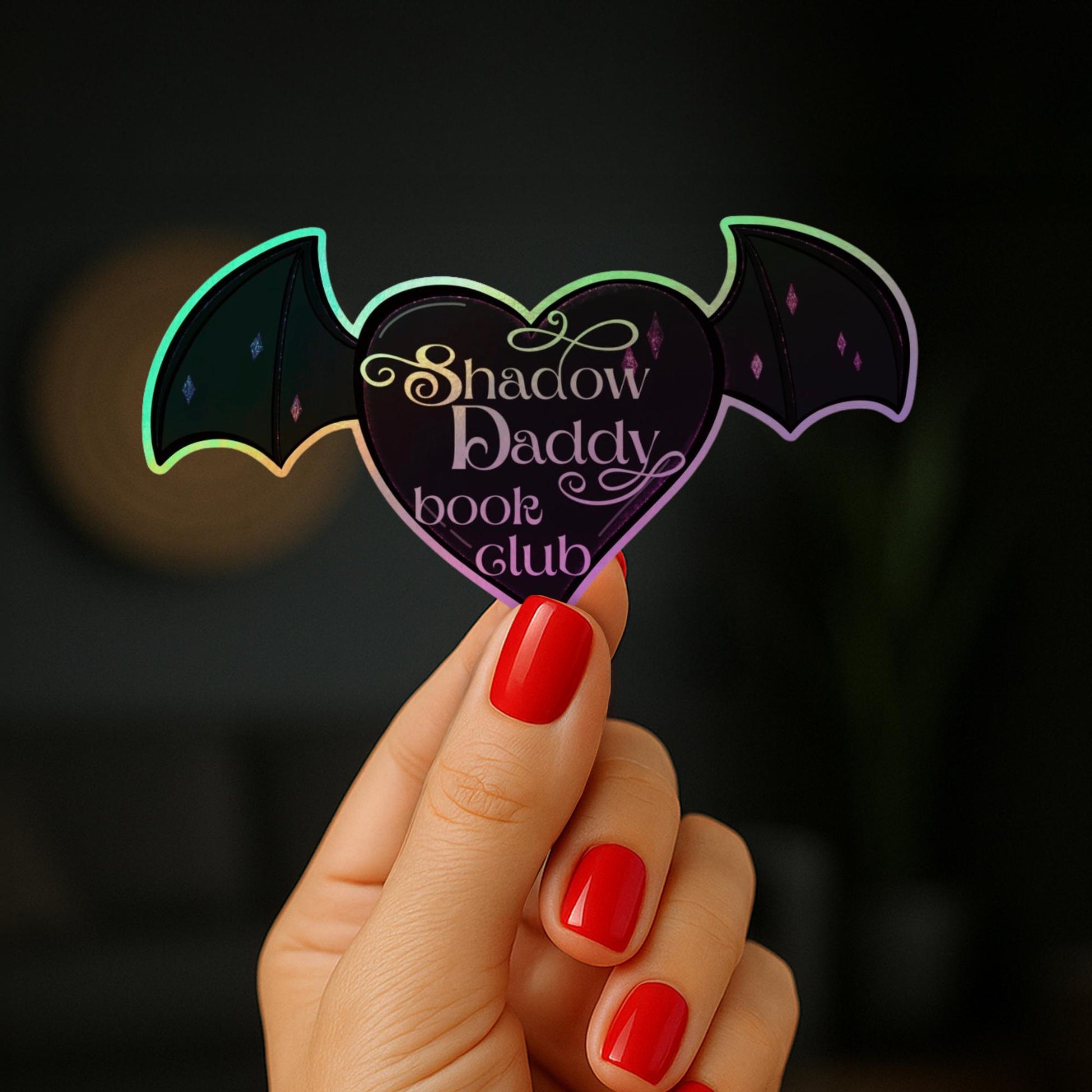 May include: A heart-shaped sticker with bat wings, featuring the text "Shadow Daddy book club." The sticker has a holographic effect with iridescent colours. The text is in a decorative script font.