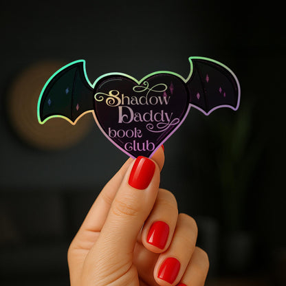 May include: A heart-shaped sticker with bat wings, featuring the text "Shadow Daddy book club." The sticker has a holographic effect with iridescent colours. The text is in a decorative script font.