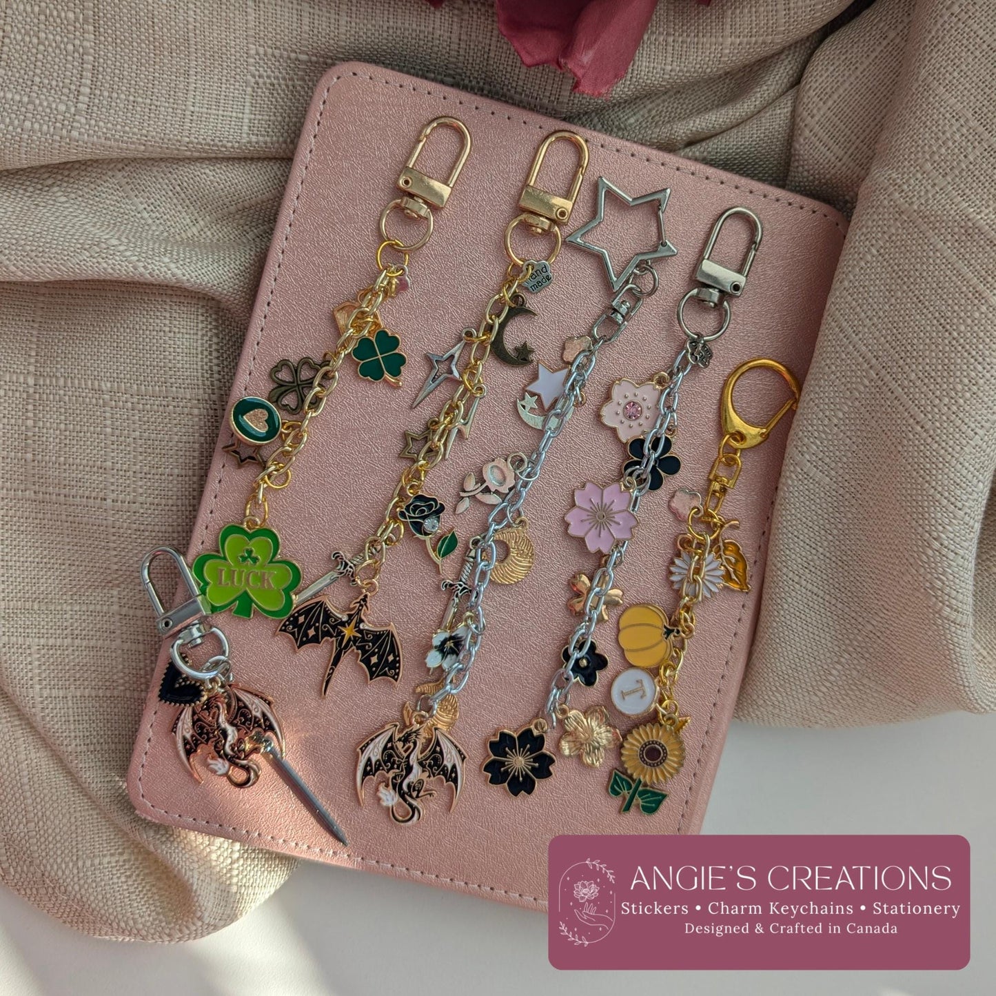 Five hand assembled romantasy and bookish charm keychains displayed on a pink notebook, featuring dragon charm, four leaf clover, celestial moon, floral and fantasy inspired designs in gold silver and black hardware, waterproof and durable, perfect Kindle eReader accessory and book lover gift, designed and crafted in Ottawa Canada by Angie's Creations