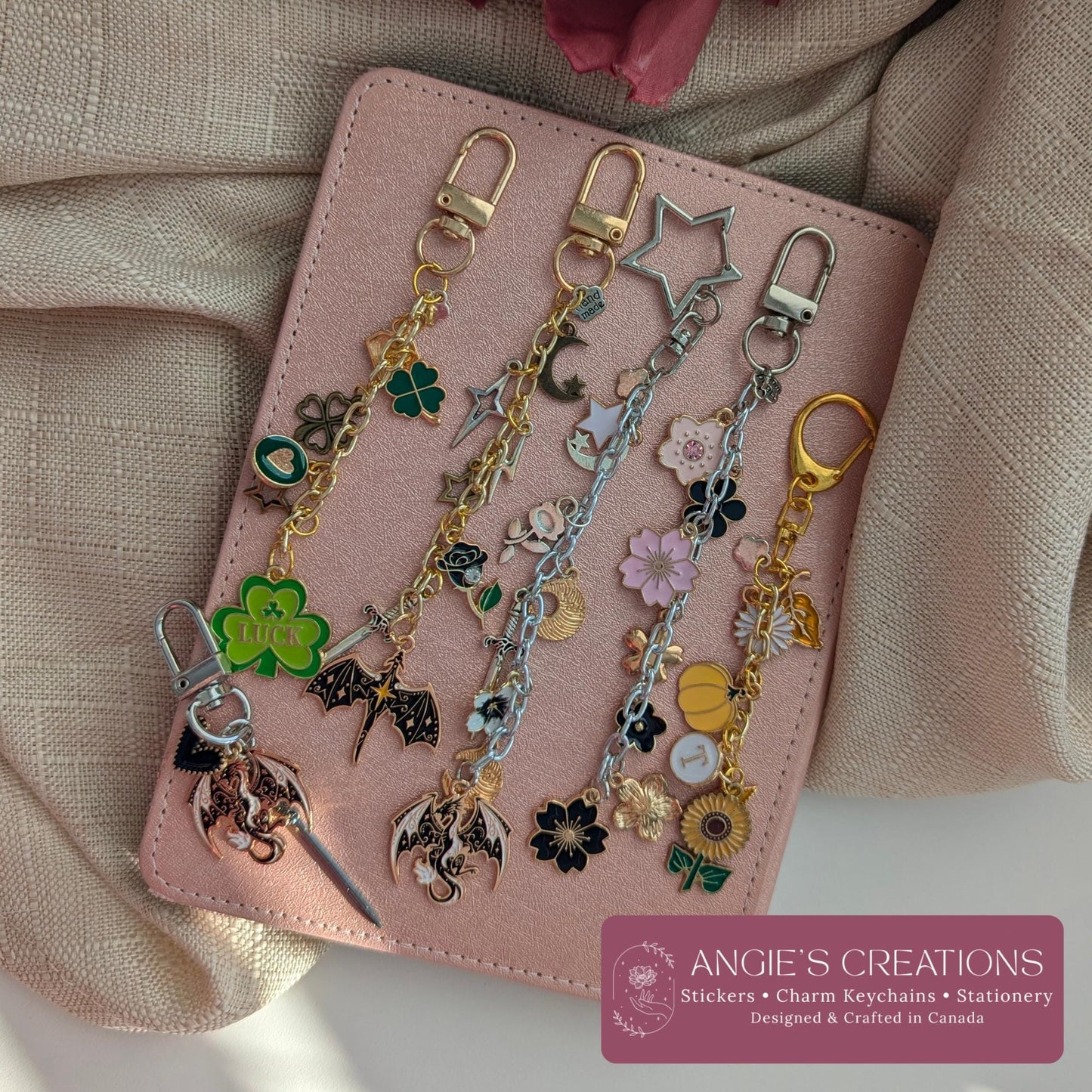 Five hand assembled romantasy and bookish charm keychains displayed on a pink notebook, featuring dragon charm, four leaf clover, celestial moon, floral and fantasy inspired designs in gold silver and black hardware, waterproof and durable, perfect Kindle eReader accessory and book lover gift, designed and crafted in Ottawa Canada by Angie's Creations
