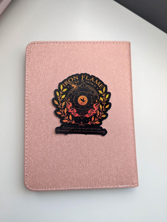 May include: A rose gold textured notebook featuring a centred sticker with the text Iron Flame Burn It Down. The sticker displays a black and orange circular emblem with a dragon silhouette, surrounded by golden laurel leaves and flames.