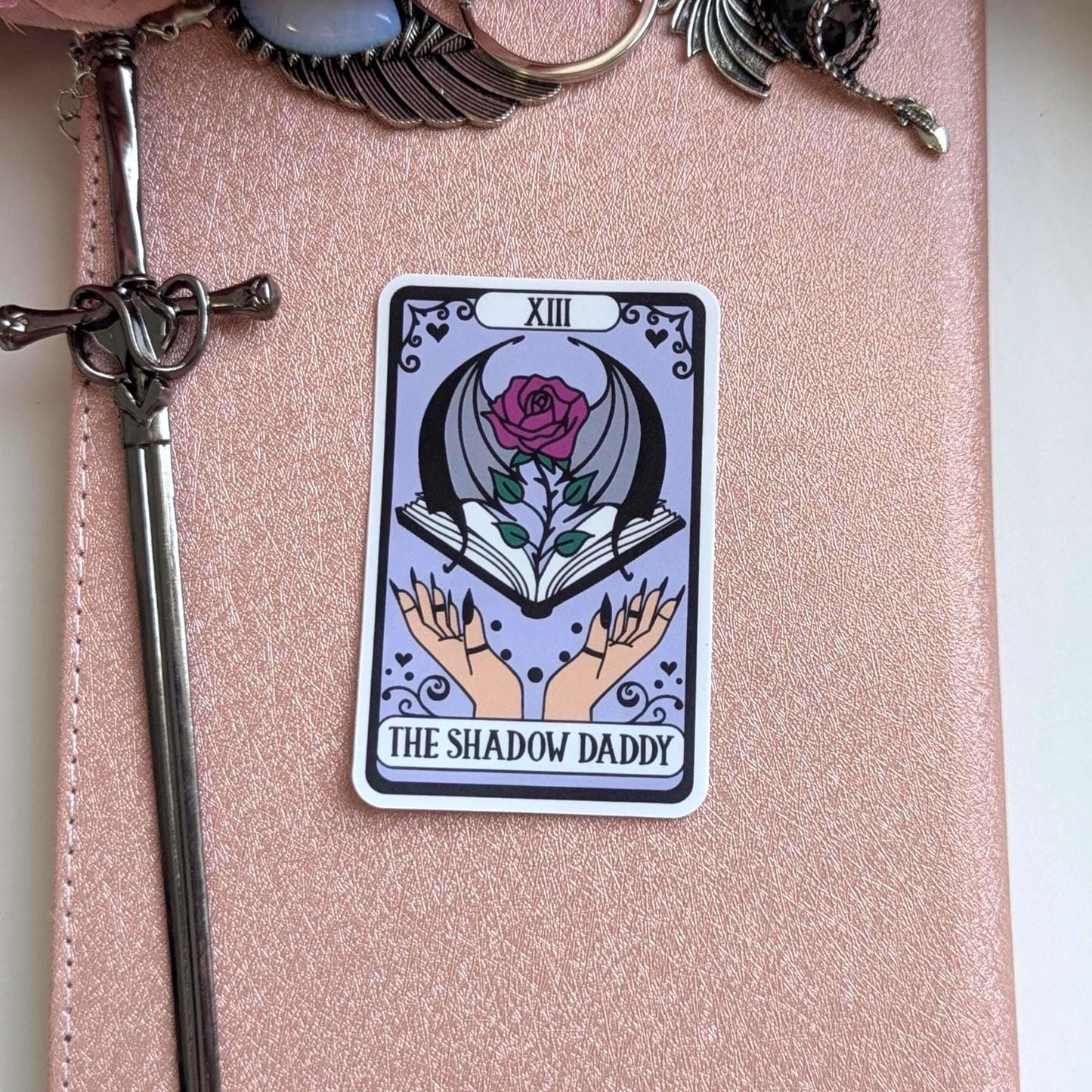 May include: A rectangular sticker featuring a tarot card design with a light purple background. The card displays the Roman numeral XIII, a rose with bat wings emerging from an open book, and two hands. The text reads THE SHADOW DADDY.