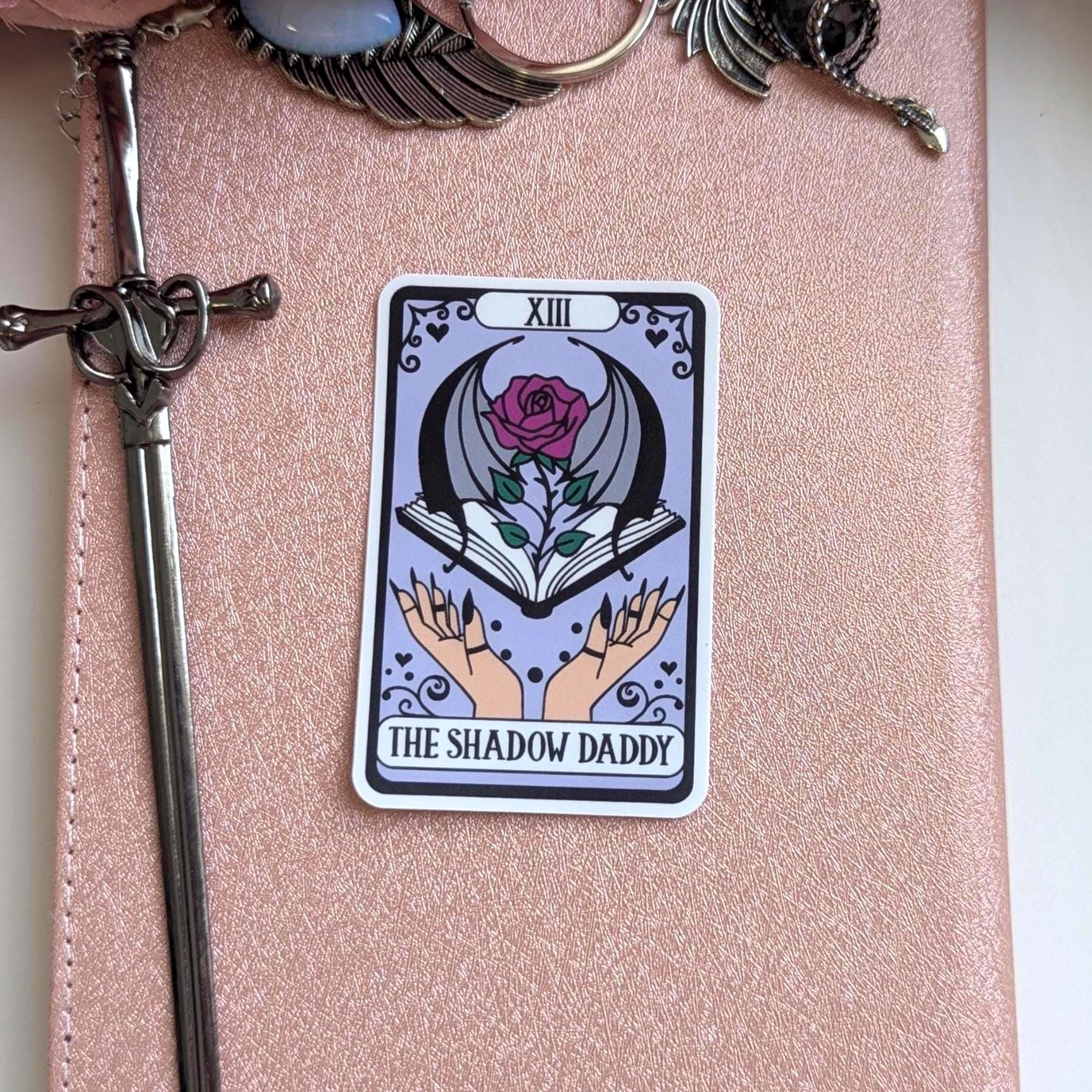 May include: A rectangular sticker featuring a tarot card design with a light purple background. The card displays the Roman numeral XIII, a rose with bat wings emerging from an open book, and two hands. The text reads THE SHADOW DADDY.