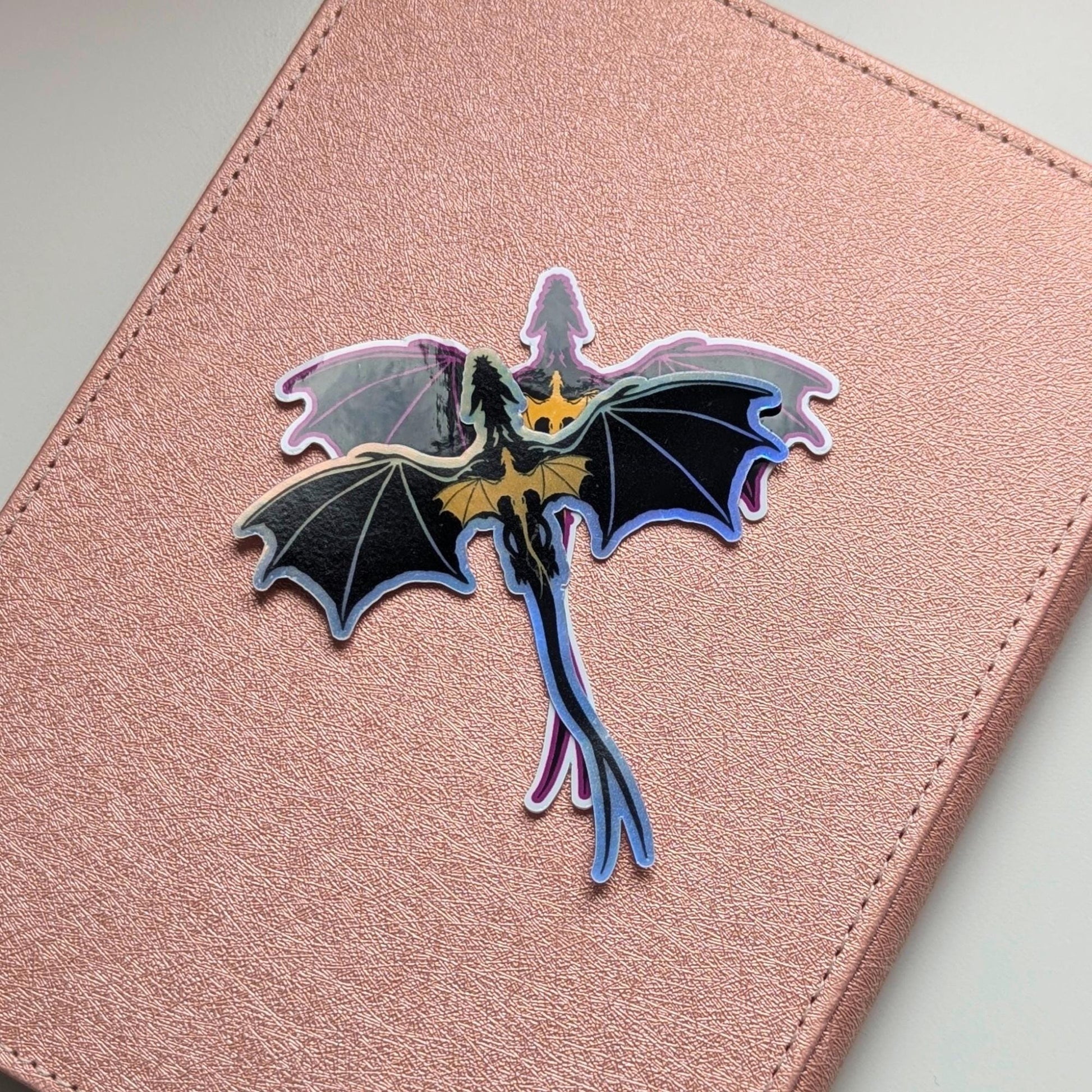 May include: A holographic dragon sticker with black wings and a gold chest, on a pink background. The dragon is depicted in flight, with a long tail. The sticker has an iridescent, shiny finish.
