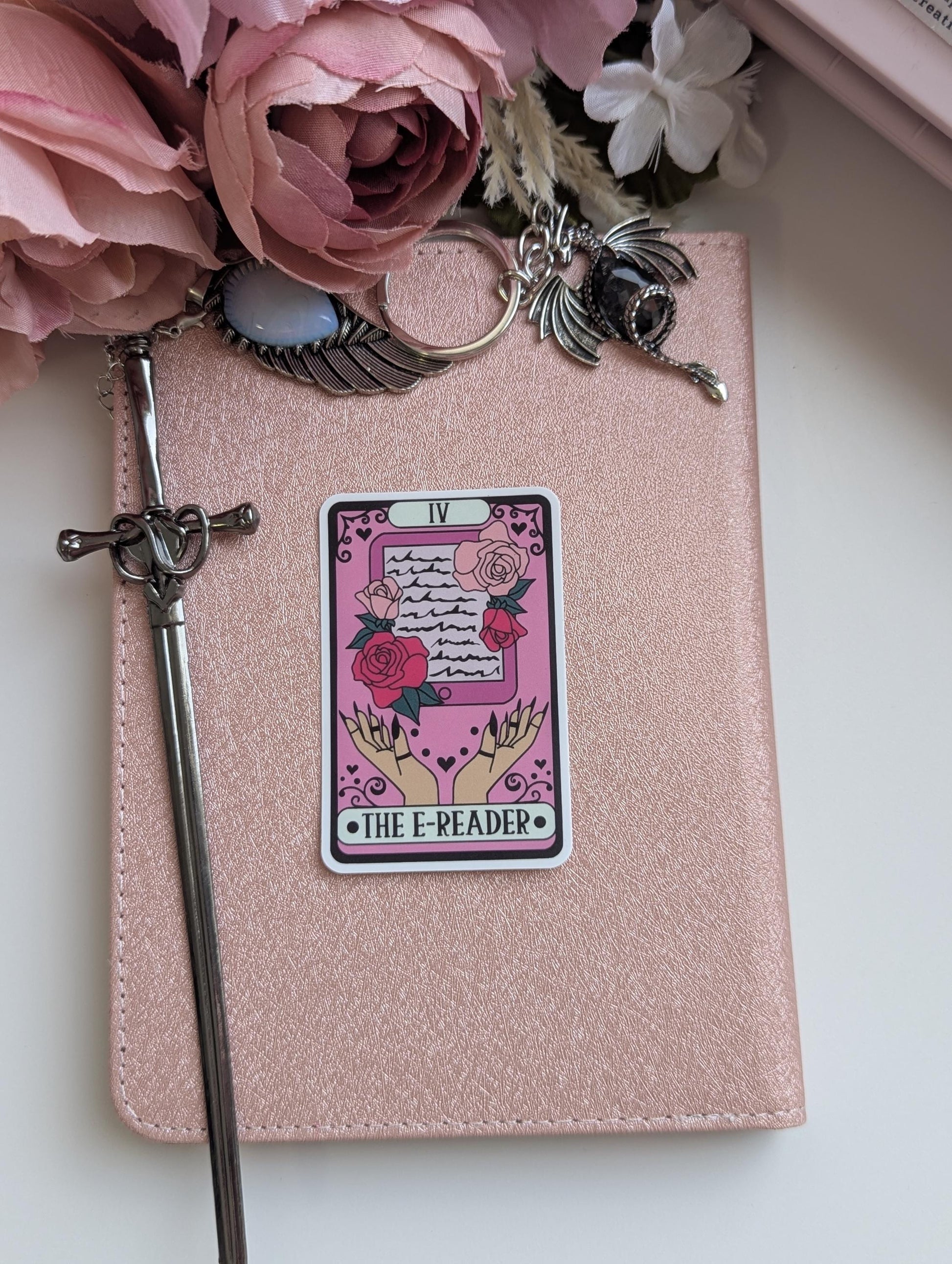 May include: A rose gold e-reader cover featuring a sticker of a tarot card labelled IV THE E-READER, depicting hands holding a tablet with roses. A silver sword-shaped stylus rests on the left, with a dragon and wing charm attached to the top.
