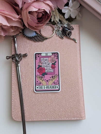 May include: A rose gold e-reader cover featuring a sticker of a tarot card labelled IV THE E-READER, depicting hands holding a tablet with roses. A silver sword-shaped stylus rests on the left, with a dragon and wing charm attached to the top.