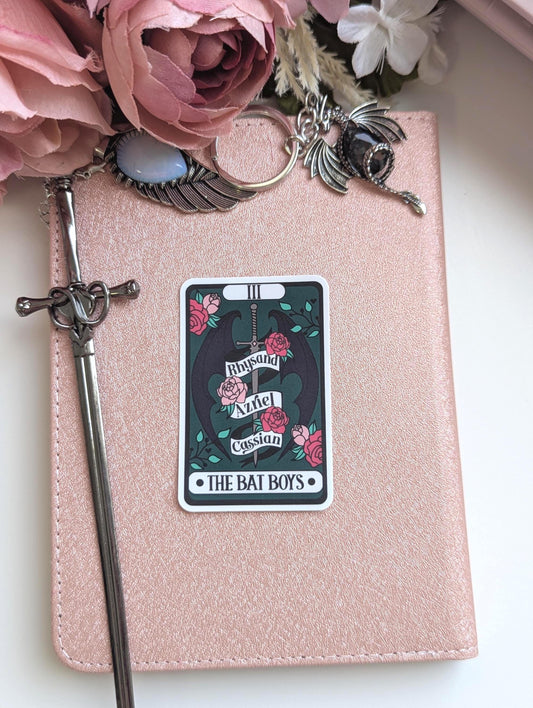 May include: A rose gold notebook featuring a rectangular sticker with a dark green background, a central sword, black wings, and pink roses. The sticker displays the text Rhysand, Azriel, Cassian, and THE BAT BOYS. A silver sword bookmark and dragon charm are attached.