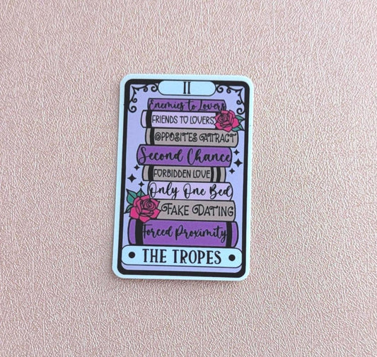 May include: A rectangular sticker designed like a tarot card featuring a stack of books in shades of purple and grey. The books are labelled with text including Enemies to Lovers, Friends to Lovers, Opposites Attract, Second Chance, Forbidden Love, Only One Bed, Fake Dating, and Forced Proximity, with pink roses and the text THE TROPES.