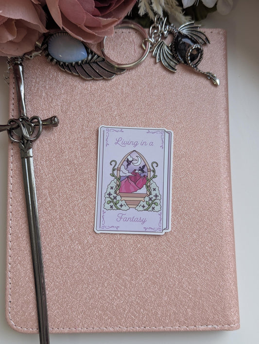 May include: A rose gold notebook featuring a rectangular sticker with the text Living in a Fantasy, depicting a mountain landscape through an archway. A silver sword-shaped bookmark, a wing charm with a blue stone, and a dragon charm are attached.