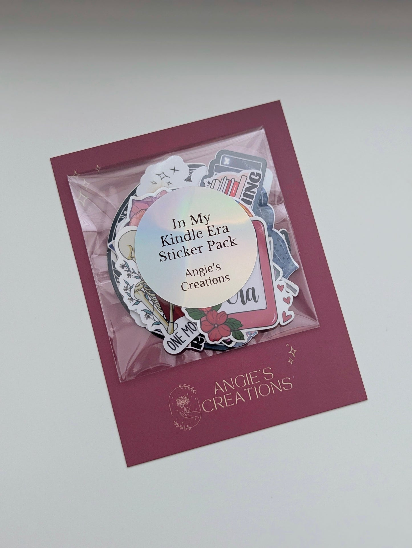 Angie's Creations "In My Kindle Era" holographic bookish sticker pack displayed in a clear resealable bag on a dark rose branded backing card, showing a collection of mixed die-cut stickers including romantasy, dragon, and reader-themed designs with the Angie's Creations logo. Handmade bookish sticker bundle designed in Ottawa Canada, perfect for Kindle cases, laptops, water bottles, journals, and planners.